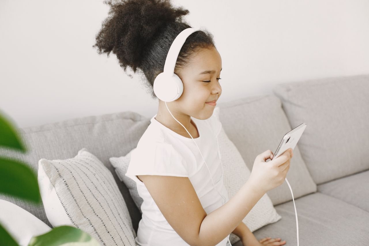Happy child sitting on sofa, listening to music with headphones and smartphone indoors.